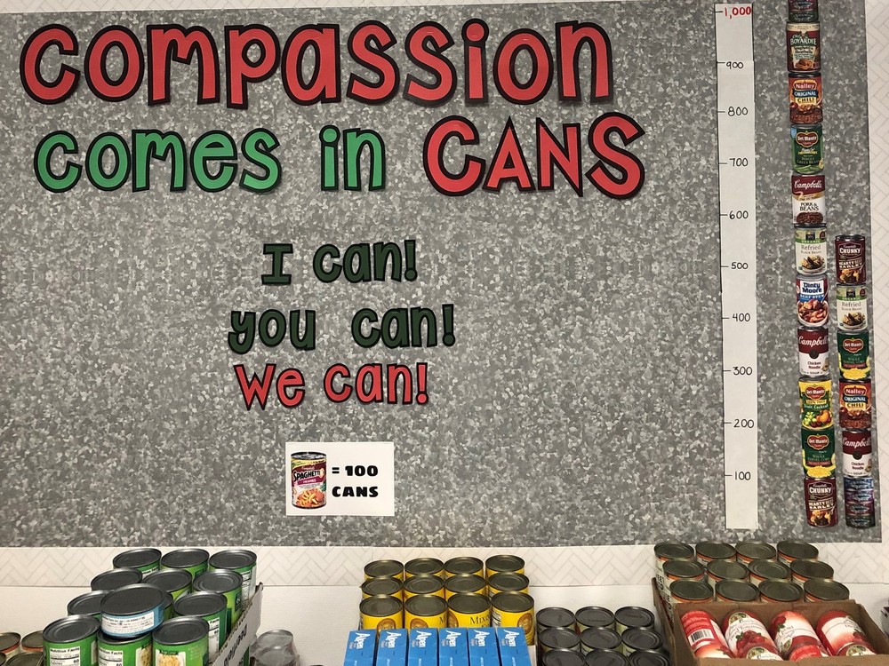 Bulletin board that says, "Compassion comes in cans. I can! You can! We can!" cans of food are piled below the board