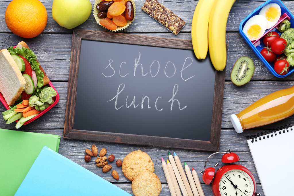 Chalk board that reads school lunch, fruits and vegetables, pencils, a clock, notepad, sandwich, orange, bananas